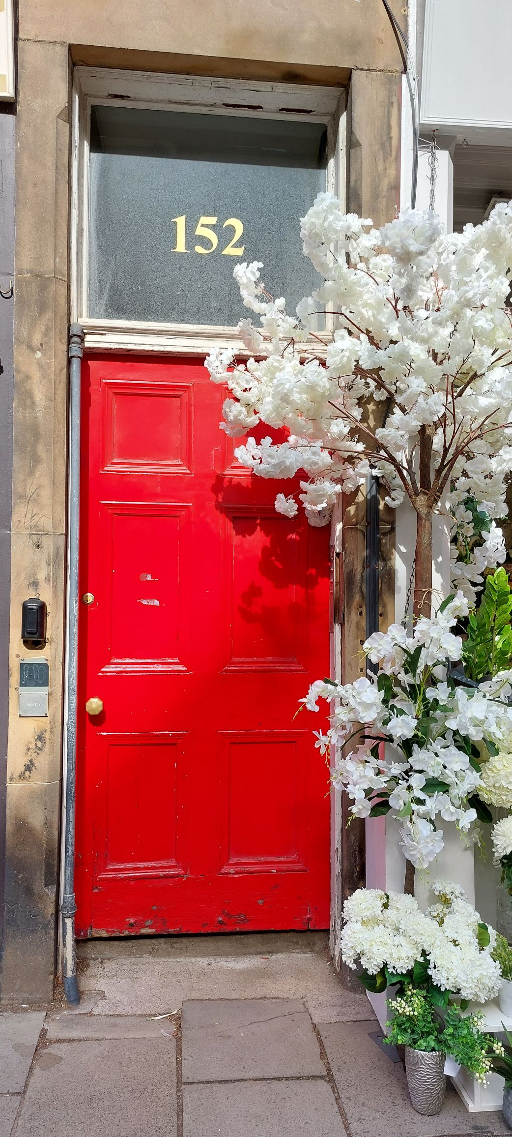 A vibrant red door numbered 152 is framed by elegant white floral arrangements. The door features traditional paneling and a large window above, enhancing the inviting entrance of the building.