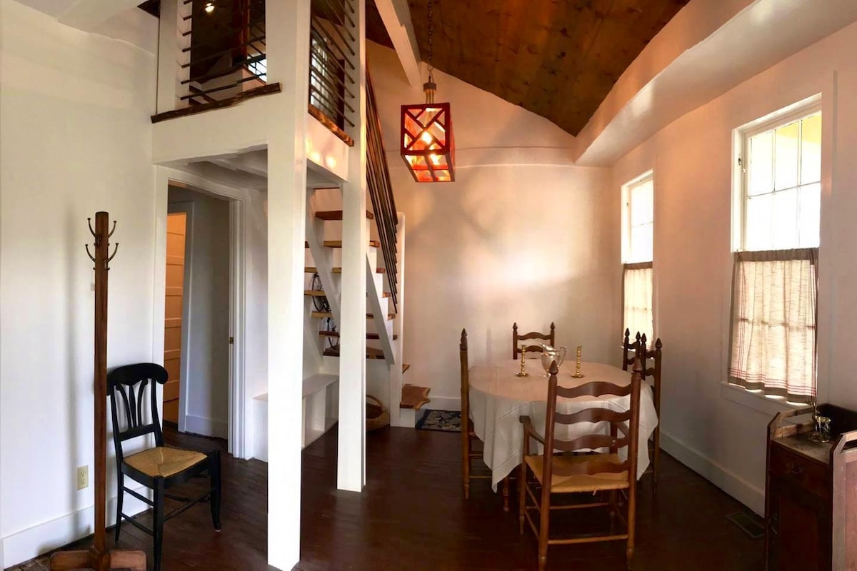 A dining area is illuminated by a unique light fixture hanging from a high ceiling. A round table set for dining is surrounded by wooden chairs. Stairs lead to an upper level, and windows allow natural light to enhance the space.