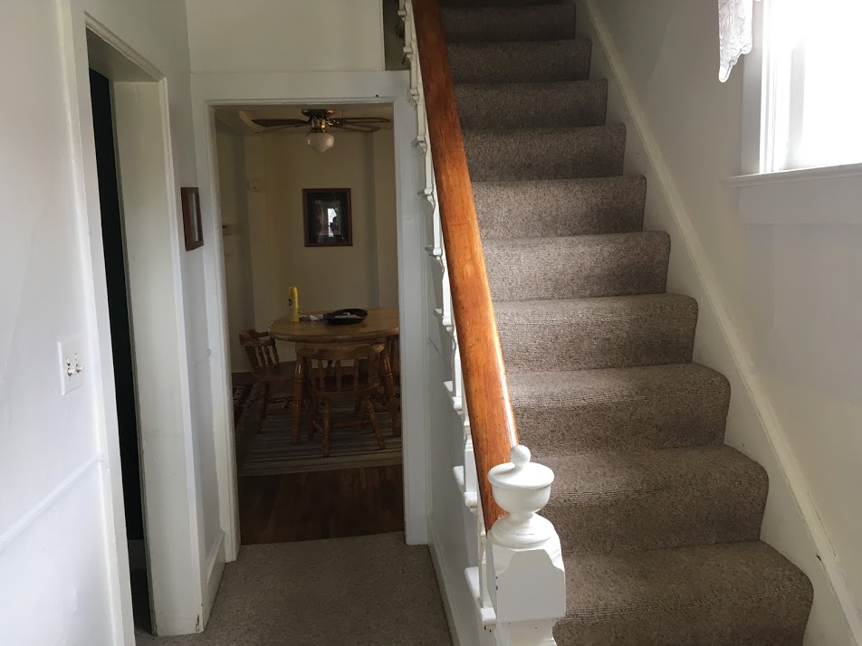 A carpeted staircase leads to the second floor, featuring a wooden handrail that complements the light-colored walls. A doorway at the end of the hallway provides a view into the dining area, where a table and chairs are visible in a well-lit space.