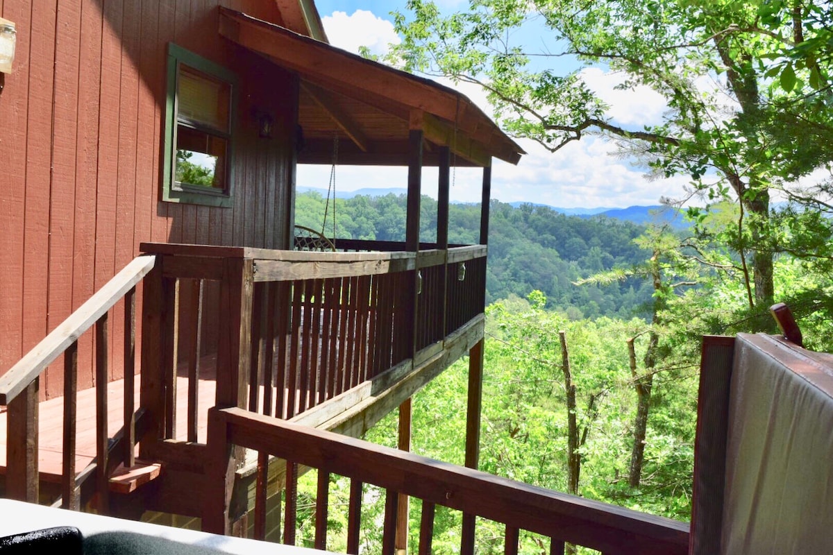 A wooden deck extends from the cabin, surrounded by lush greenery. The view reveals rolling hills and forested landscapes bathed in natural light, with an inviting swing hanging from the porch. The structure's rustic red exterior complements the serene mountain setting.