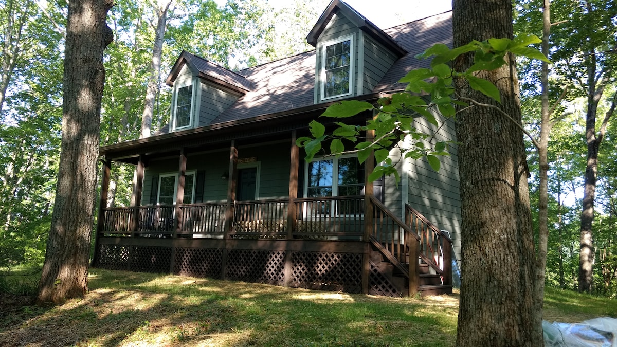 A spacious cabin is nestled among trees, featuring a welcoming front porch supported by sturdy columns. Large windows allow natural light to enter, while a grassy area surrounds the cabin, enhancing the sense of privacy and seclusion.