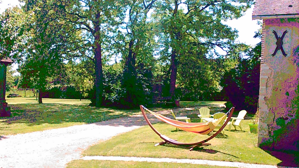 A serene outdoor area is highlighted by a gently swaying hammock and several light-colored chairs arranged on a grassy space. Mature trees provide ample shade, creating a peaceful environment. The gravel path leads towards a charming stone structure, enhancing the tranquil countryside setting.