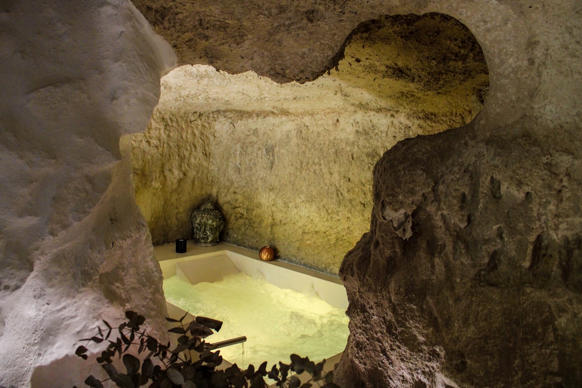 An intimate space is created within the cave, featuring a private, climate-controlled jacuzzi nestled against the rocky walls. Soft illumination highlights the natural textures of the stone, while a serene atmosphere is enhanced by decorative elements placed nearby.