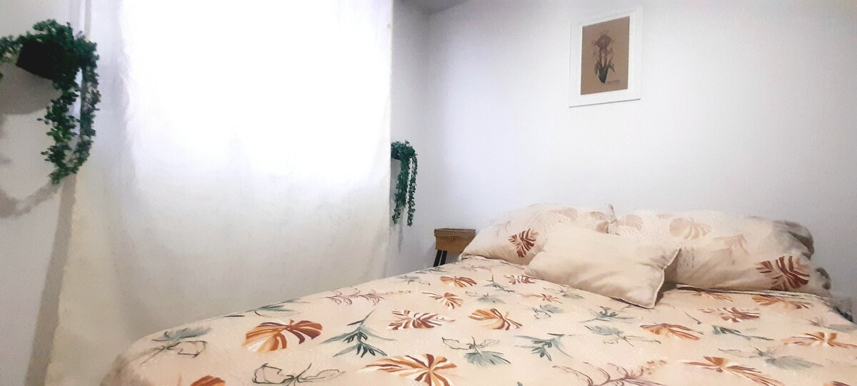 A cozy sleeping area features a spacious bed adorned with a floral-patterned duvet and soft pillows. Natural light filters through a large window, complemented by minimalist decor and greenery on the walls, creating a serene environment.