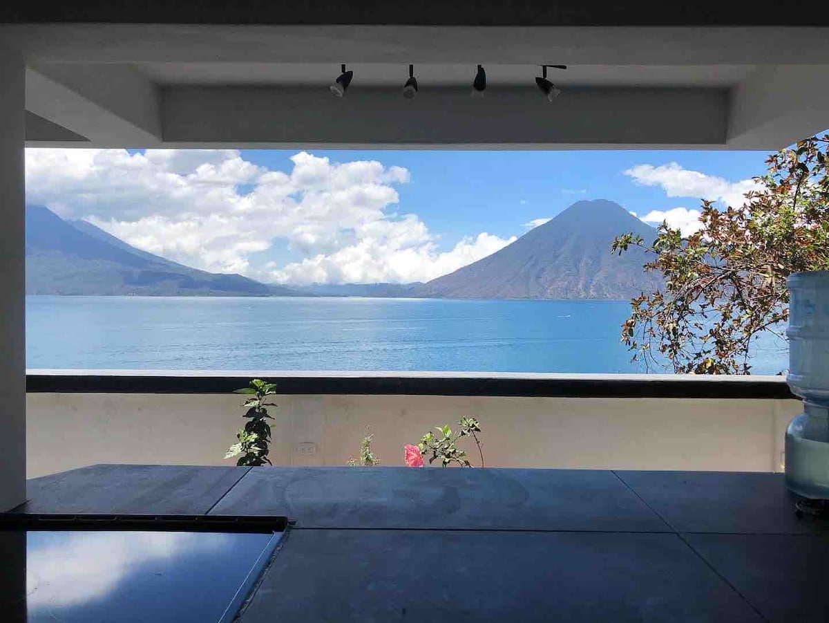 A view of Lake Atitlan is visible from a kitchen space, framed by a large opening that merges indoor and outdoor elements. Mountains surround the lake, with vibrant greenery in the foreground. Natural light enhances the serene atmosphere.
