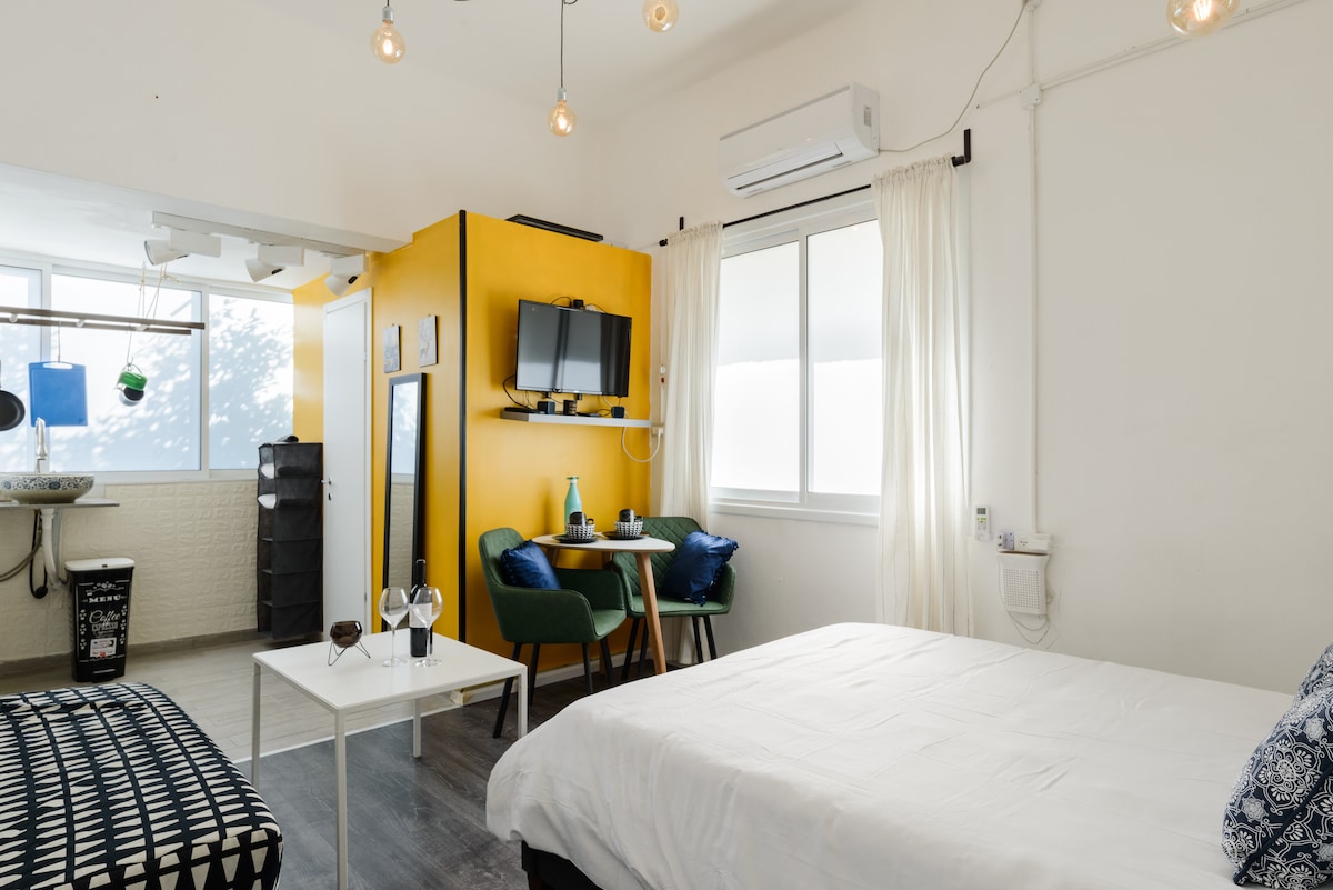 A bright studio apartment features a spacious double bed and a cozy seating area with green chairs. A yellow accent wall supports a TV, and natural light enters through large windows with sheer curtains. A well-equipped kitchen area and modern furnishings are also visible.