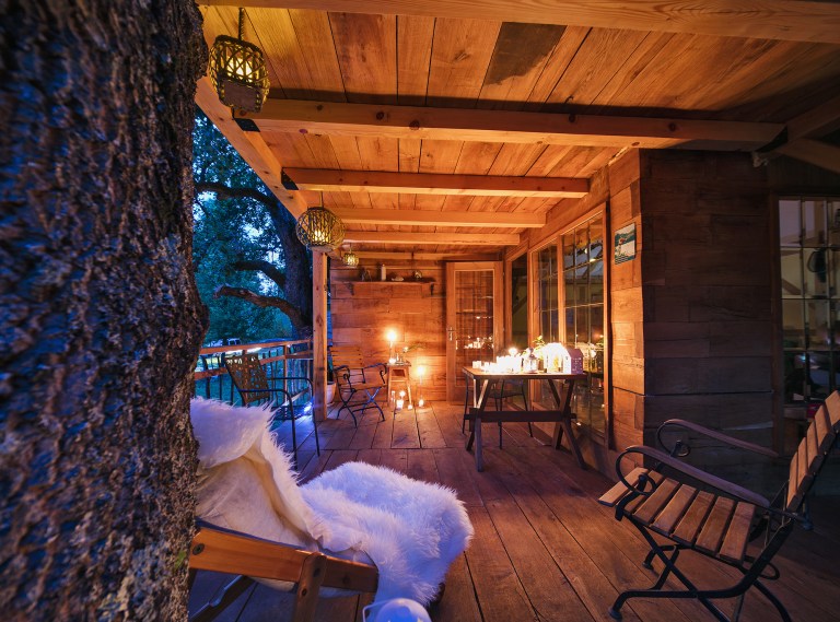 An inviting outdoor deck is framed by warm wooden beams, featuring comfortable seating arrangements. Soft lighting illuminates a table set for dining, while textured throws and lanterns add a cozy touch. Large glass doors offer views of the surrounding natural landscape.