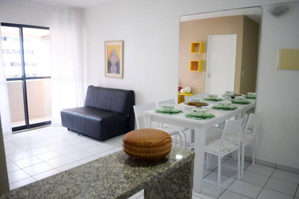 Balcão de apoio para a cozinha ou apoio para trabalhos, mesa jantar com 4 cadeiras, sofá cama que acomoda mais duas pessoas com ventilador de teto, varanda com vista para a avenida principal.