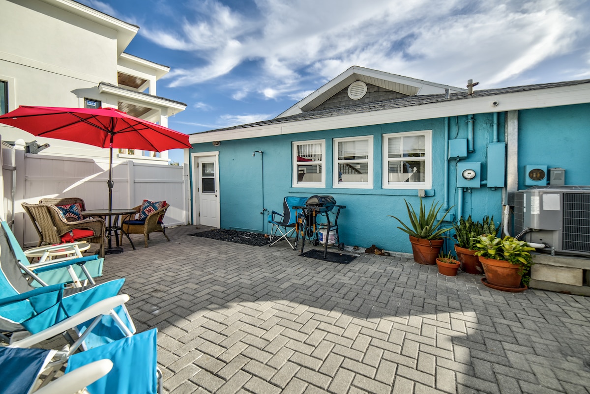 An outdoor patio area features colorful blue and white walls. A red umbrella provides shade over a seating arrangement with chairs and a small table. Potted plants add greenery, while a grill is positioned nearby. The space is paved with stone and enclosed by a white fence.