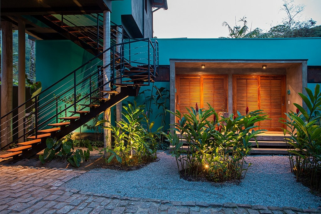 The exterior entrance is framed by lush greenery, with strategically placed lighting highlighting the plants. A staircase leads to the upper level, while two wooden doors serve as the main entry point, set against a vibrant blue wall.
