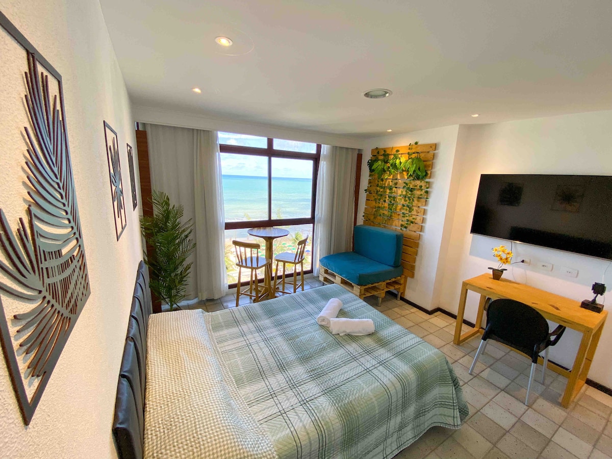 The room features a spacious bed covered with a checkered blanket, complemented by a wall-mounted TV. A work desk and chair are positioned near a large window, providing views of the beach. A small dining table with seating is visible beside a blue lounge area.