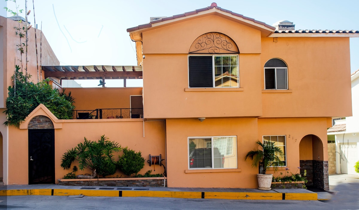 The exterior of a two-story home is depicted, featuring warm orange walls, decorative arches above the windows, and a balcony area adorned with greenery. A spacious driveway is visible, along with a landscaped area of low-maintenance plants near the main entrance.