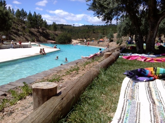 A natural swimming area is visible, featuring a clear blue pool bordered by sandy edges. Guests can be seen enjoying the water, while colorful blankets are spread on the green grass nearby. Lush trees and a hilly landscape frame the serene setting.