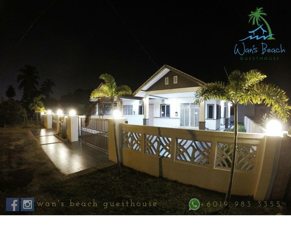 The exterior of the guesthouse is illuminated at night, showcasing the light-colored building and its welcoming entrance. Decorative palm trees line the pathway, and the secure fence features a modern design with stone accents, enhancing the overall appeal of the property.