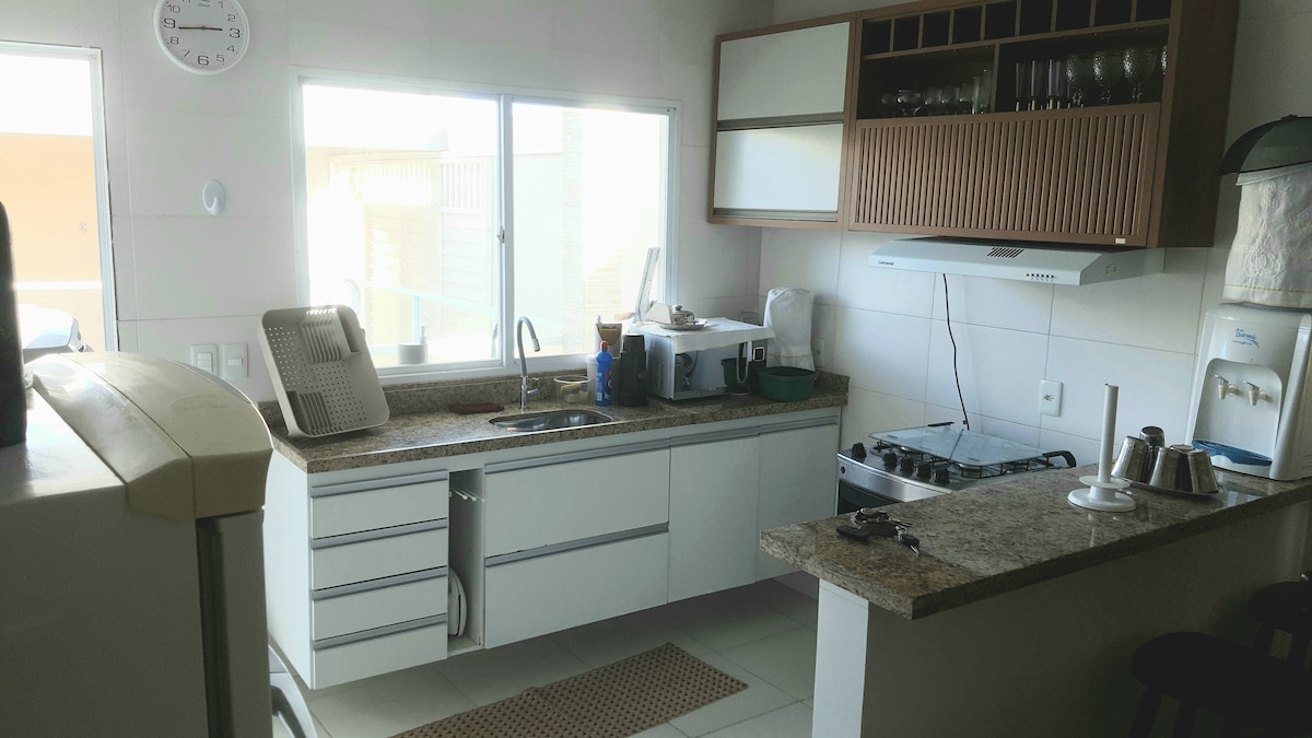 A modern kitchen features a spacious countertop with sleek cabinets above. The kitchen is equipped with a stove, sink, and various appliances, including a refrigerator. Natural light fills the space through a large window, enhancing the functional layout.