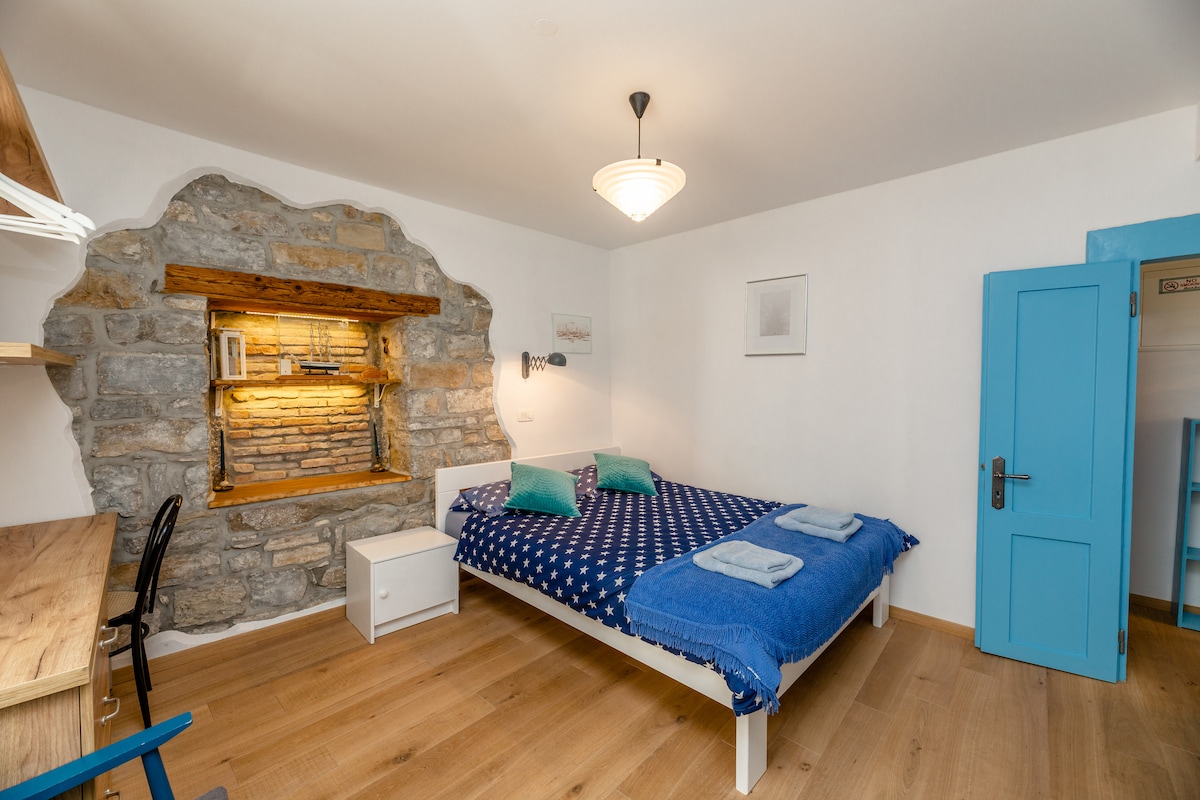 A spacious sleeping area features a stone wall with a built-in niche, illuminated softly. A large bed with a navy blue duvet and pillows invites rest. A light wood bedside table and a desk with a chair sit nearby, enhancing the room's functional layout.
