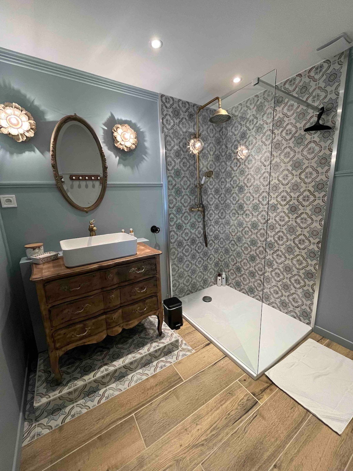 A modern bathroom features a glass shower enclosure with a rainfall showerhead. A vintage-style wooden vanity with a white basin sits against a patterned wall. Soft lighting fixtures and a round mirror enhance the space, while textured flooring adds warmth.