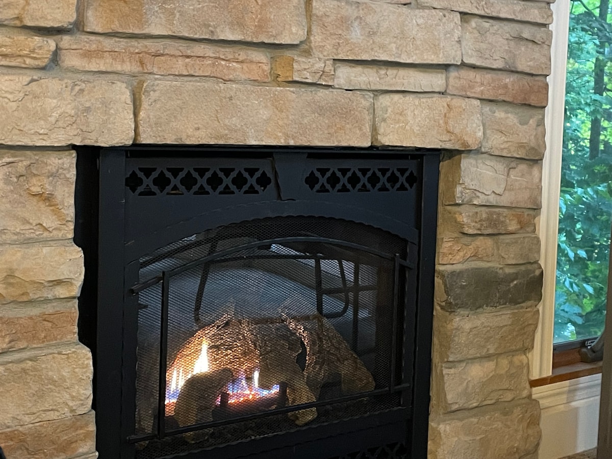 A stone fireplace is accented by natural stonework in warm tones, featuring a gas flame that flickers above arranged logs. A view of greenery is visible through a nearby window, creating a cozy atmosphere.