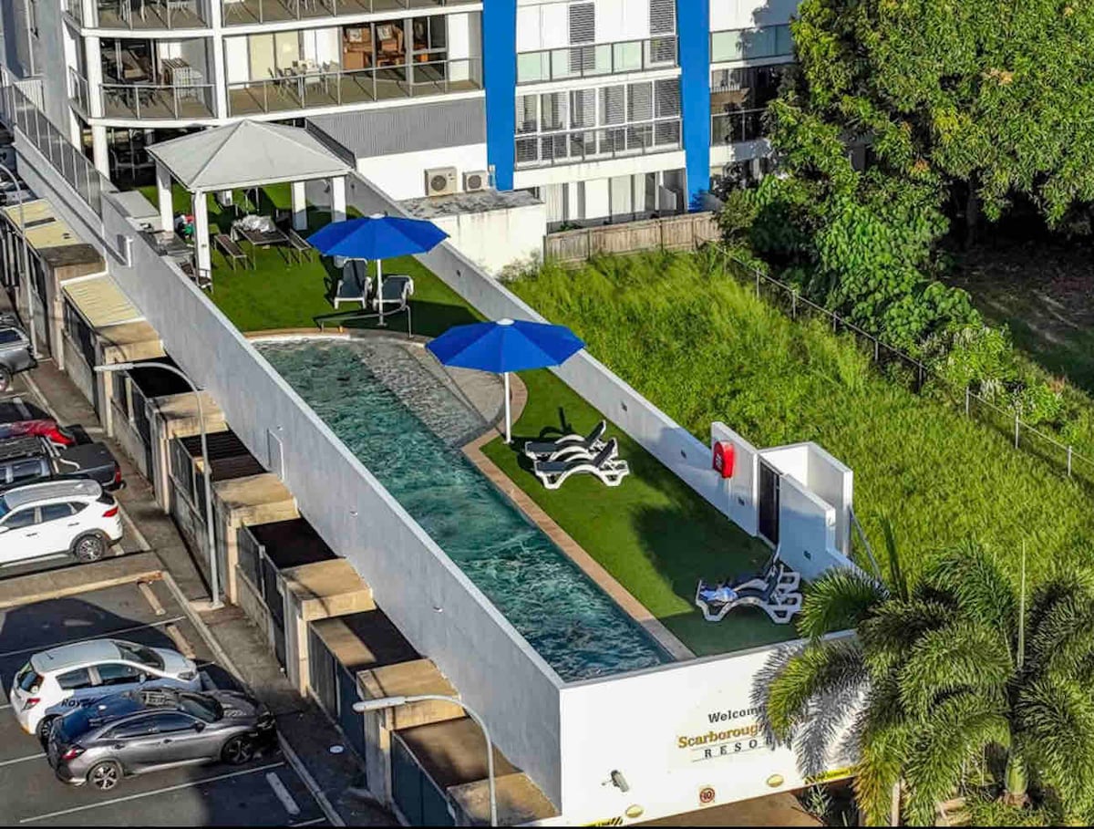 An aerial view showcases a serene pool area lined with sun loungers and large umbrellas. The pool is surrounded by well-maintained grass, enhancing the outdoor space. Nearby, a covered seating area provides shade, contributing to a relaxing environment in the complex.