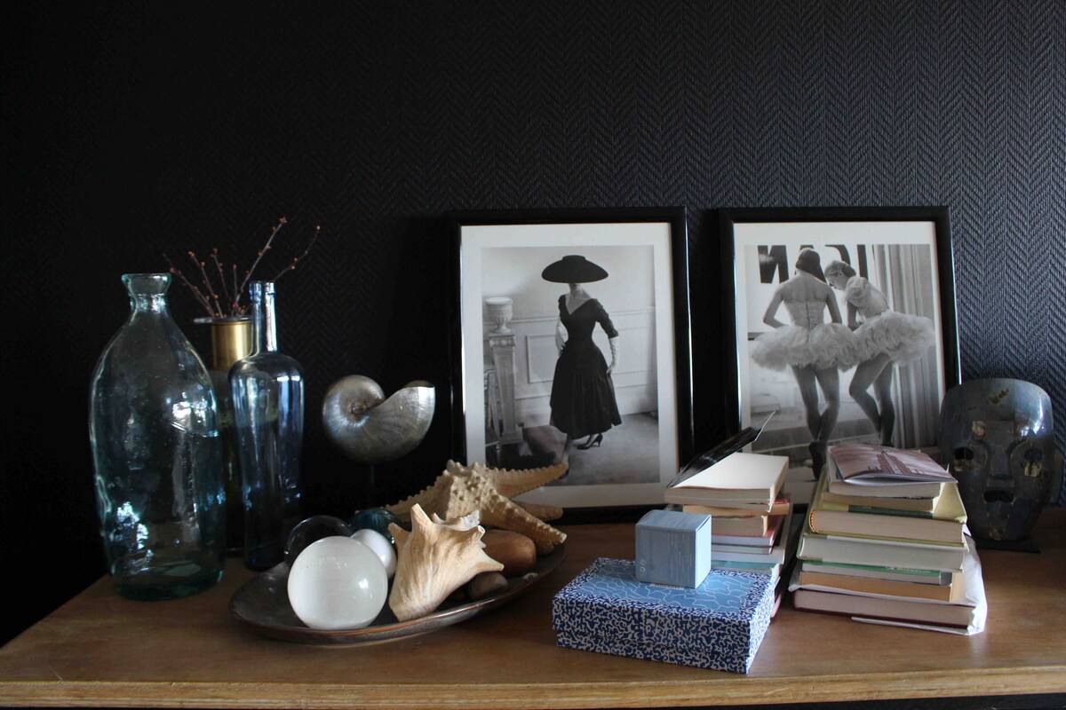 A wooden table displays artful arrangements, including two framed photographs of figures in elegant attire. Various decorative items such as glass vases, shells, and stacked books add to the overall aesthetic, with a textured black wall serving as a backdrop.