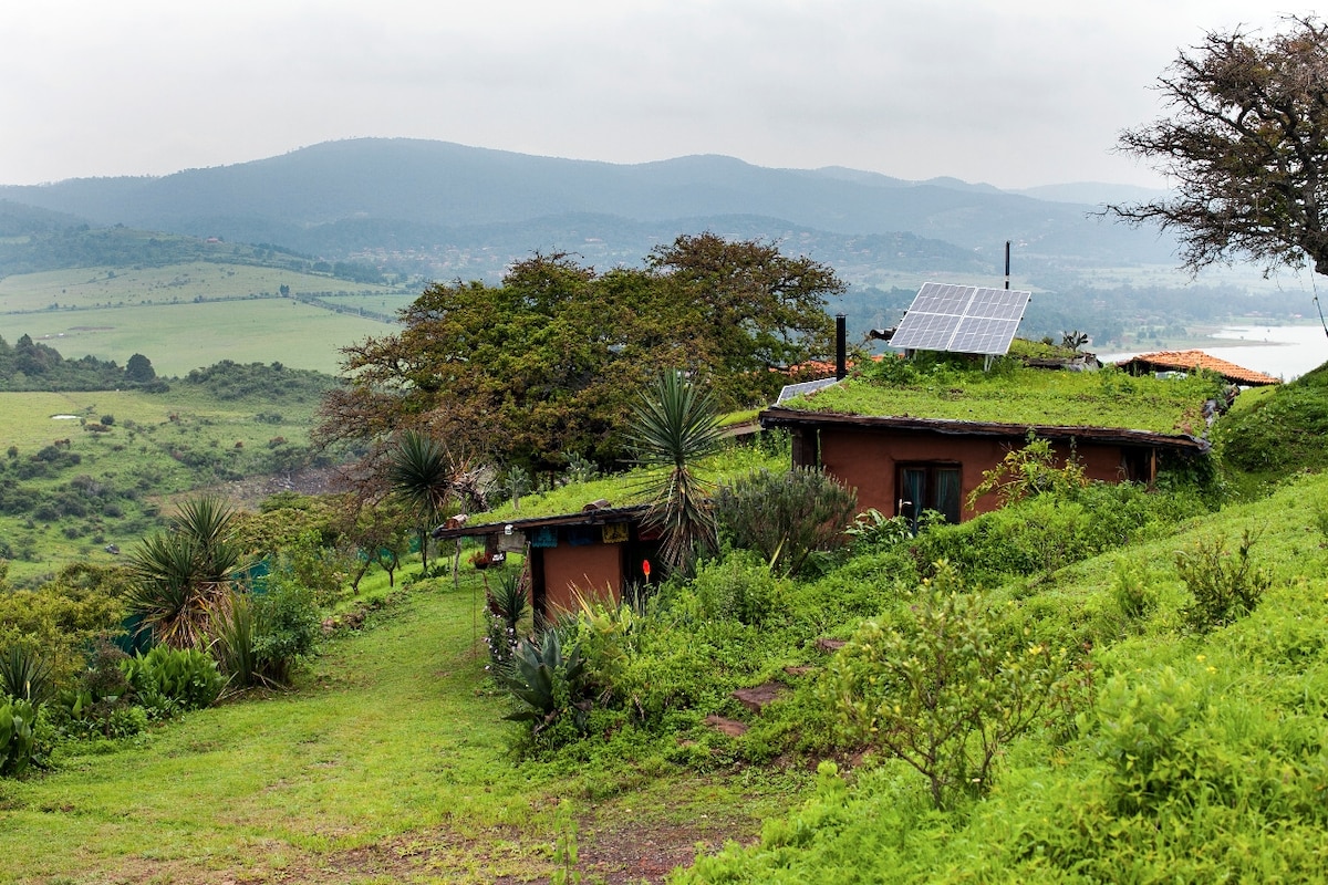 A rustic alternative construction is nestled among lush greenery, featuring a green roof that blends seamlessly with the landscape. Solar panels are visible atop the structure, contributing to sustainable energy use. Scenic hills and distant mountains provide a serene backdrop, enhancing the natural surroundings.