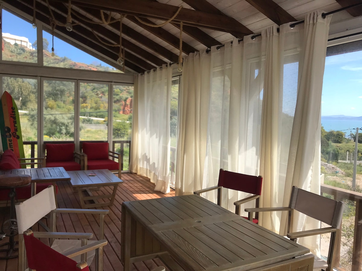 A sunlit sunroom features large glass windows adorned with sheer white curtains, allowing natural light to fill the space. Wooden furniture, including a table and chairs, provides a comfortable area for relaxation, while views of the surrounding landscape can be seen through the windows.