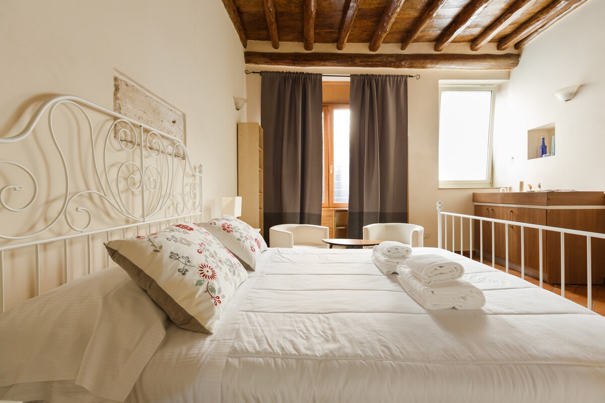 A spacious bedroom presents a comfortable bed with decorative pillows and neatly arranged towels. Natural light enters through a large window, illuminating the room. Dark curtains frame the window, and two armchairs are positioned nearby, creating a relaxing space.
