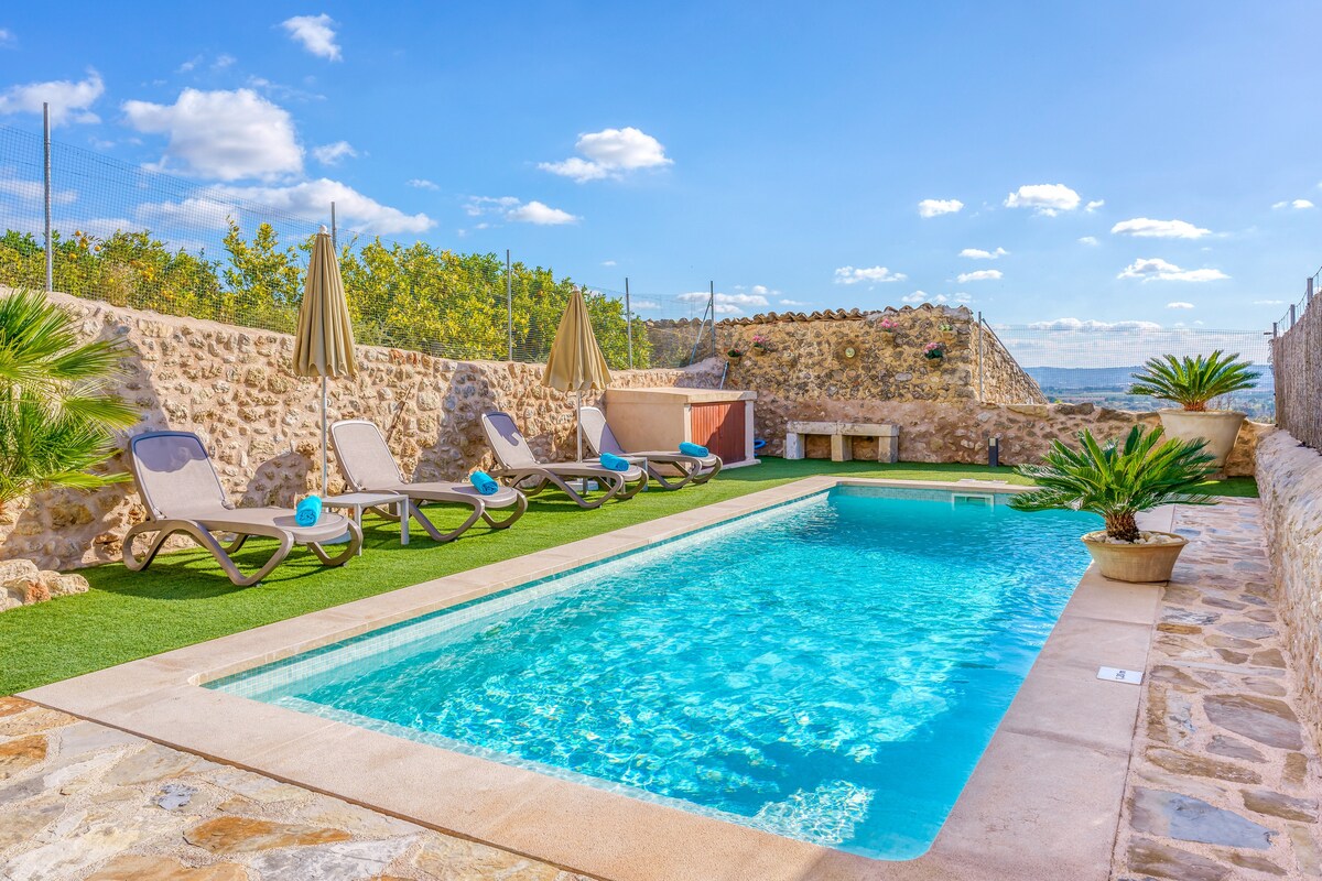 A private outdoor space features a refreshing swimming pool surrounded by sun loungers and umbrella shades. Lush greenery enhances the setting, while a stone wall provides privacy. The clear blue sky completes the serene atmosphere, creating an inviting space for relaxation and enjoyment.