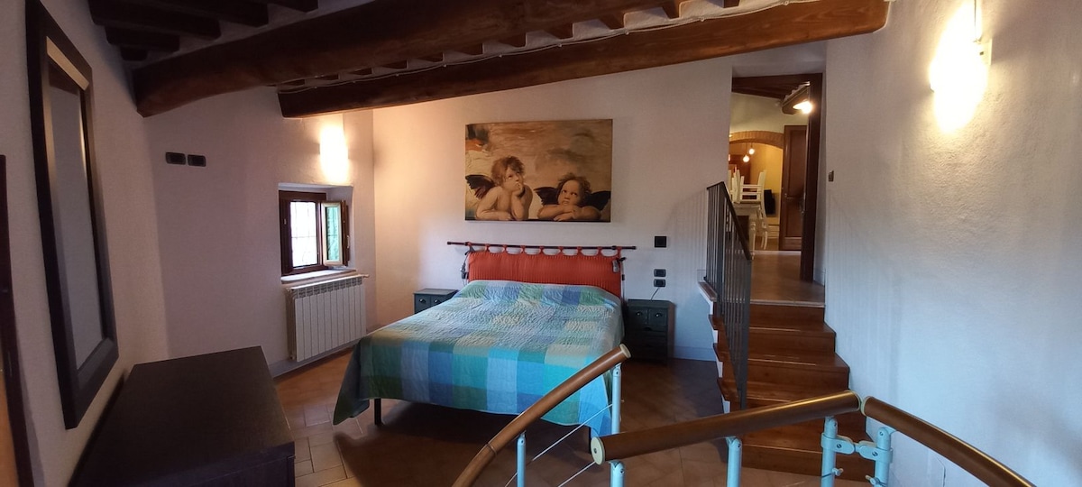 A spacious bedroom is arranged with a colorful patterned bedspread on a large bed. A piece of wall art is displayed above the headboard. Natural light enters through a window beside a radiator. A staircase leads to a separate area, adding depth to the space.