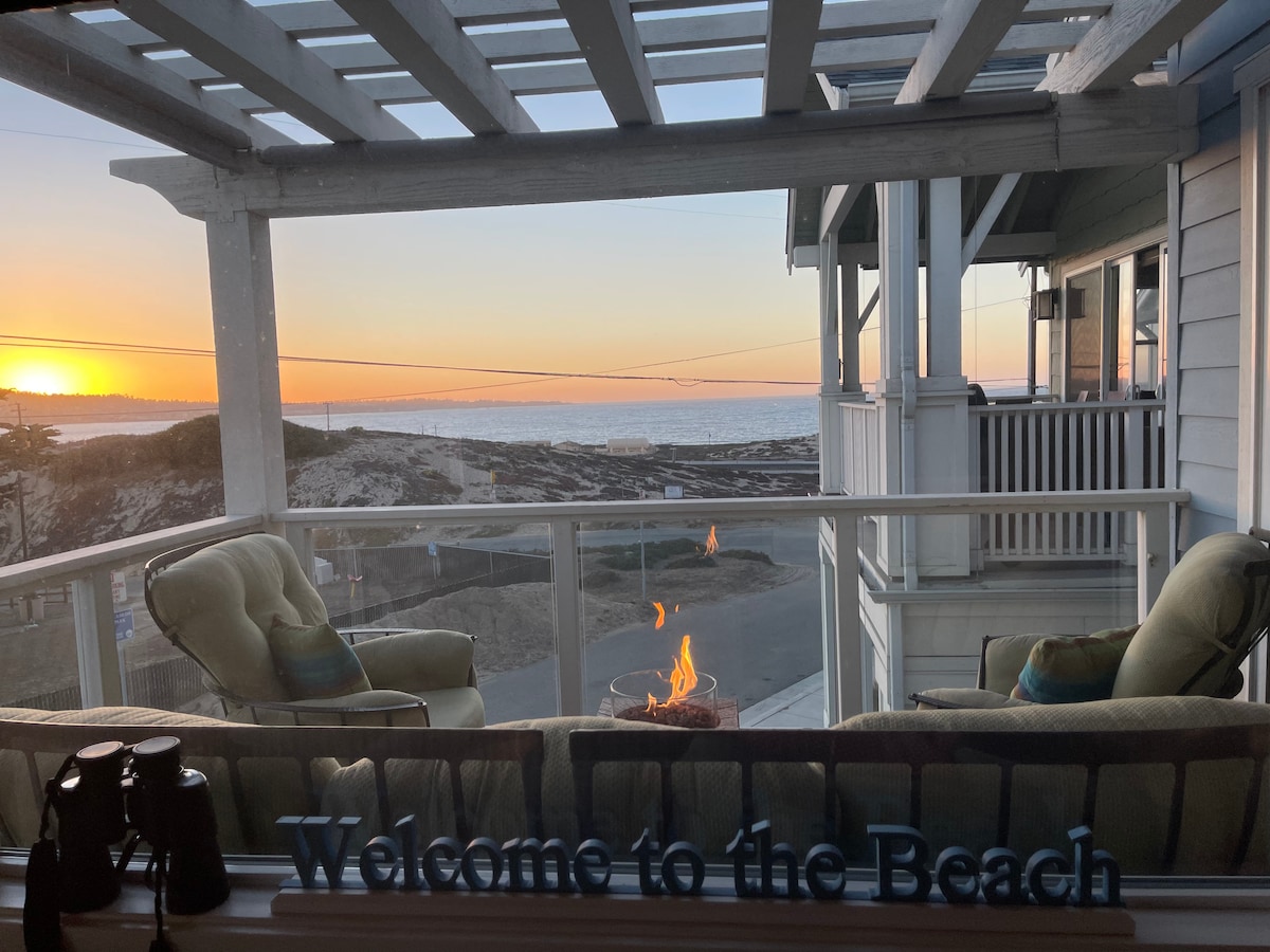 A spacious balcony provides a view of the sunset over the ocean, with a cozy seating area featuring two green chairs. A fire pit adds warmth, while a sign reading 'Welcome to the Beach' is prominently displayed in the foreground.