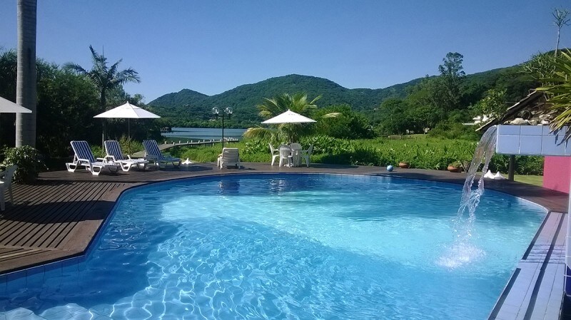 The outdoor pool is surrounded by a wooden deck, featuring lounge chairs and umbrellas for shade. Lush greenery and distant mountains provide a scenic backdrop, while a gentle waterfall feature adds a calming element to the space.
