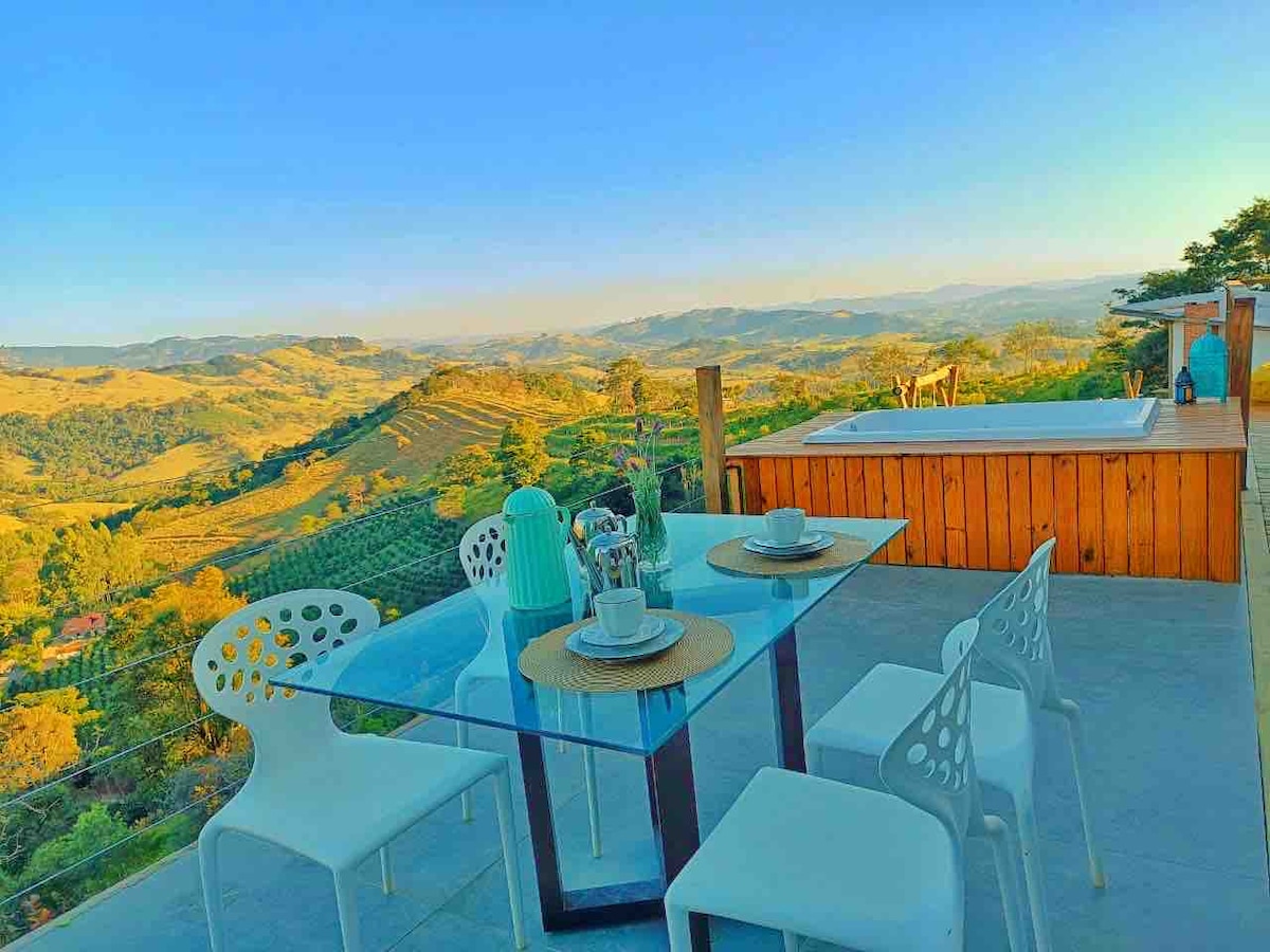 A spacious deck overlooking a stunning mountain landscape is equipped with a glass dining table and four white chairs. A jacuzzi sits nearby, inviting relaxation while taking in the breathtaking view. The scene is bathed in warm sunlight, enhancing the natural beauty surrounding the area.