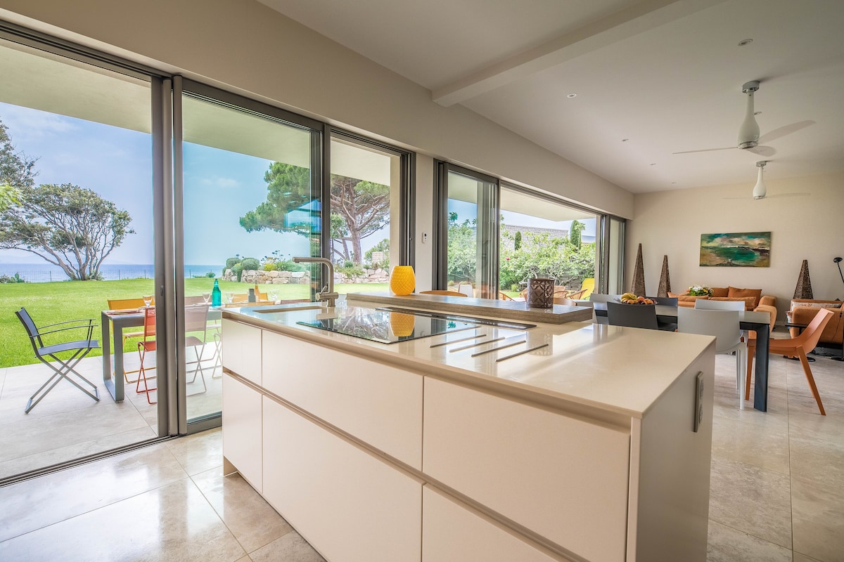 The open-concept kitchen features a large central island and is illuminated by natural light from expansive glass doors. Views of the garden and sea are visible through the sliding doors, which provide access to an outdoor dining area and lounge space.