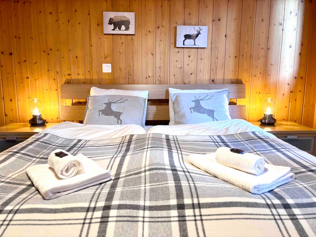 A comfortable sleeping area features a spacious bed adorned with a checked blanket and crisp white towels. Two decorative pillows with reindeer motifs complement the wood-paneled walls. Soft light from bedside lamps adds a serene ambiance to the cozy retreat.