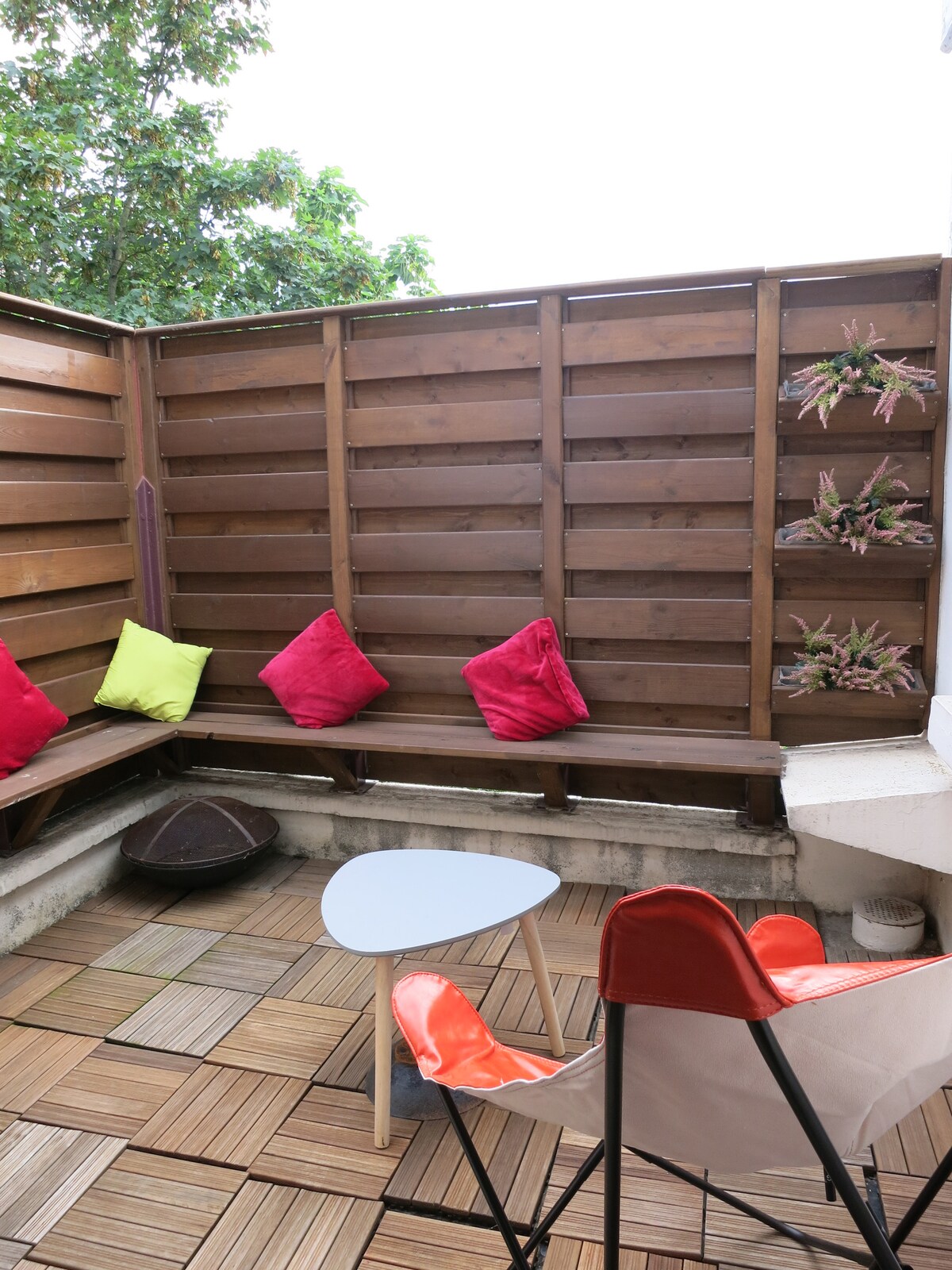 A private terrace features wooden slats for privacy and is adorned with colorful cushions in pink and lime green. A small round table is positioned alongside a comfortable chair, while potted plants enhance the space with touches of greenery.