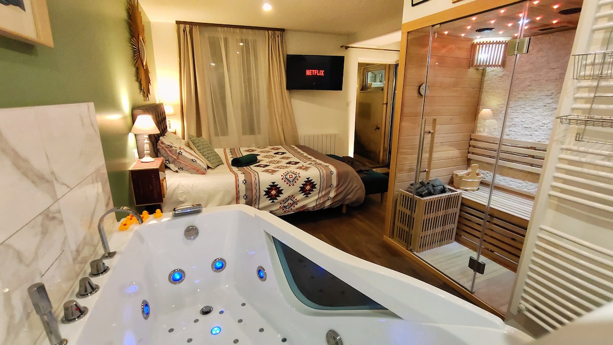 An inviting room features a spacious king-size bed with patterned bedding. A modern jacuzzi tub is positioned in the foreground, with illuminated jets. A sauna area is visible behind glass doors, alongside a shower, while a TV is mounted on the wall, enhancing the cozy atmosphere.