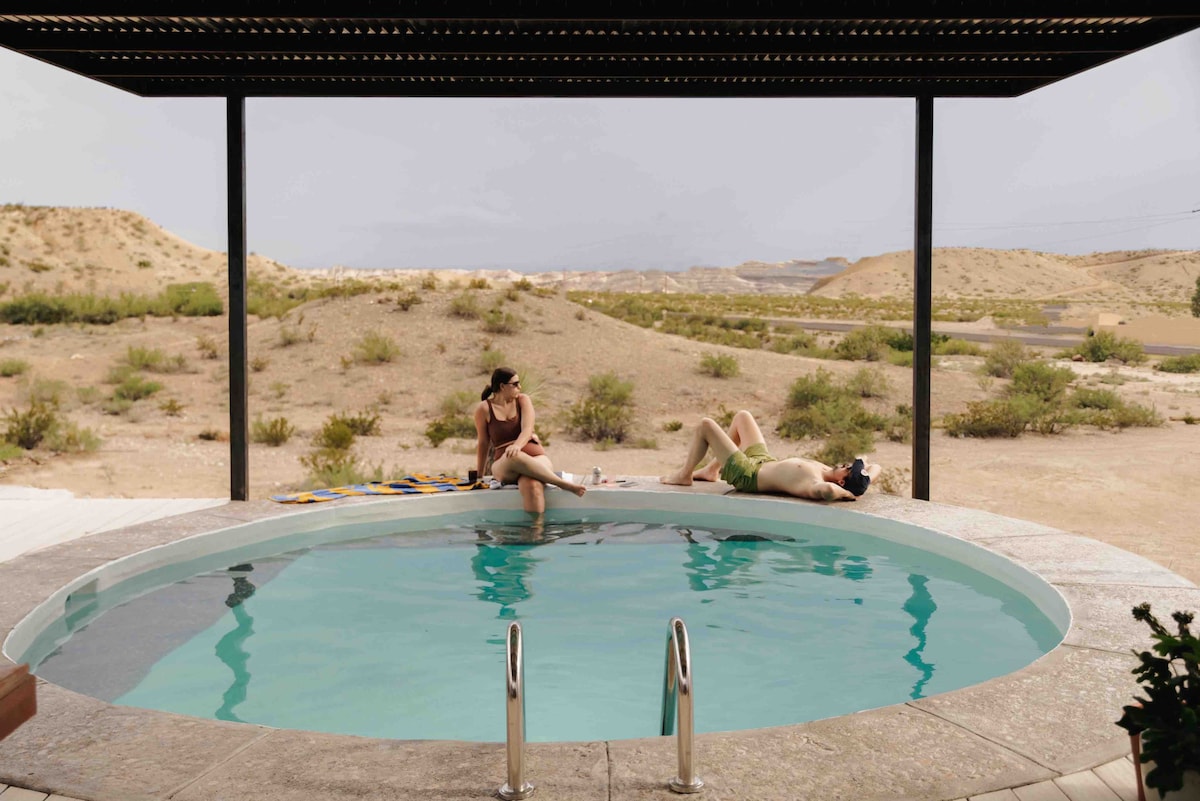 A round private pool is situated under a shaded patio, surrounded by desert landscape. Two individuals enjoy the water, while towels and drinks rest on the edge. The area is framed by two stair rails leading into the pool.