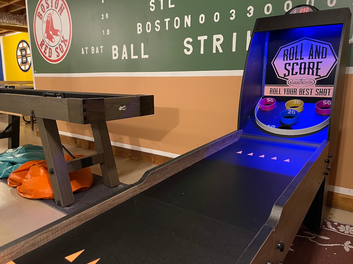 A game area features a colorful 'Roll and Score' arcade game highlighted by vibrant blue lighting. The background displays a large Boston Red Sox-themed mural, enhancing the playful atmosphere. Beside the arcade game, a shuffleboard table contributes to the entertainment options available.