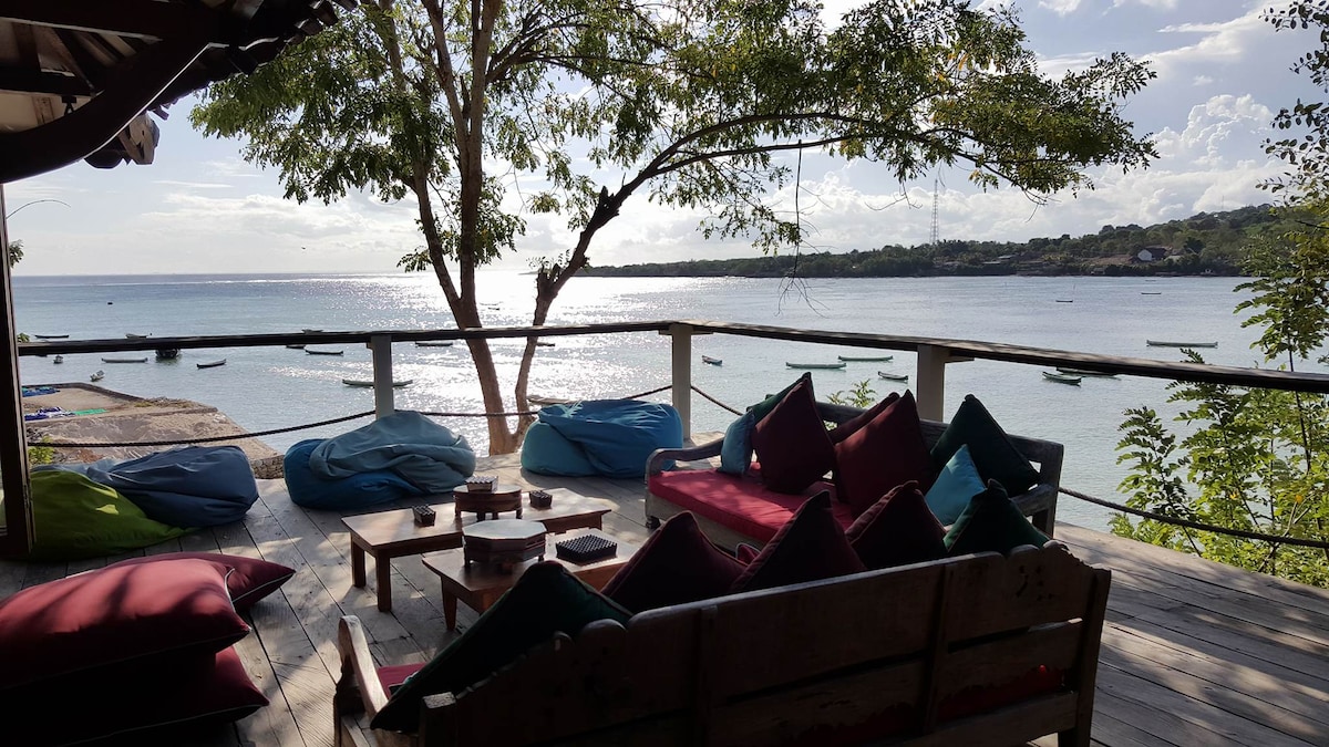 A spacious outdoor deck is furnished with colorful bean bags and wooden seating, providing a comfortable area to relax. The shimmering lagoon can be seen in the background, reflecting the clouds and sunlight. Lush greenery frames the scene, enhancing the natural beauty.