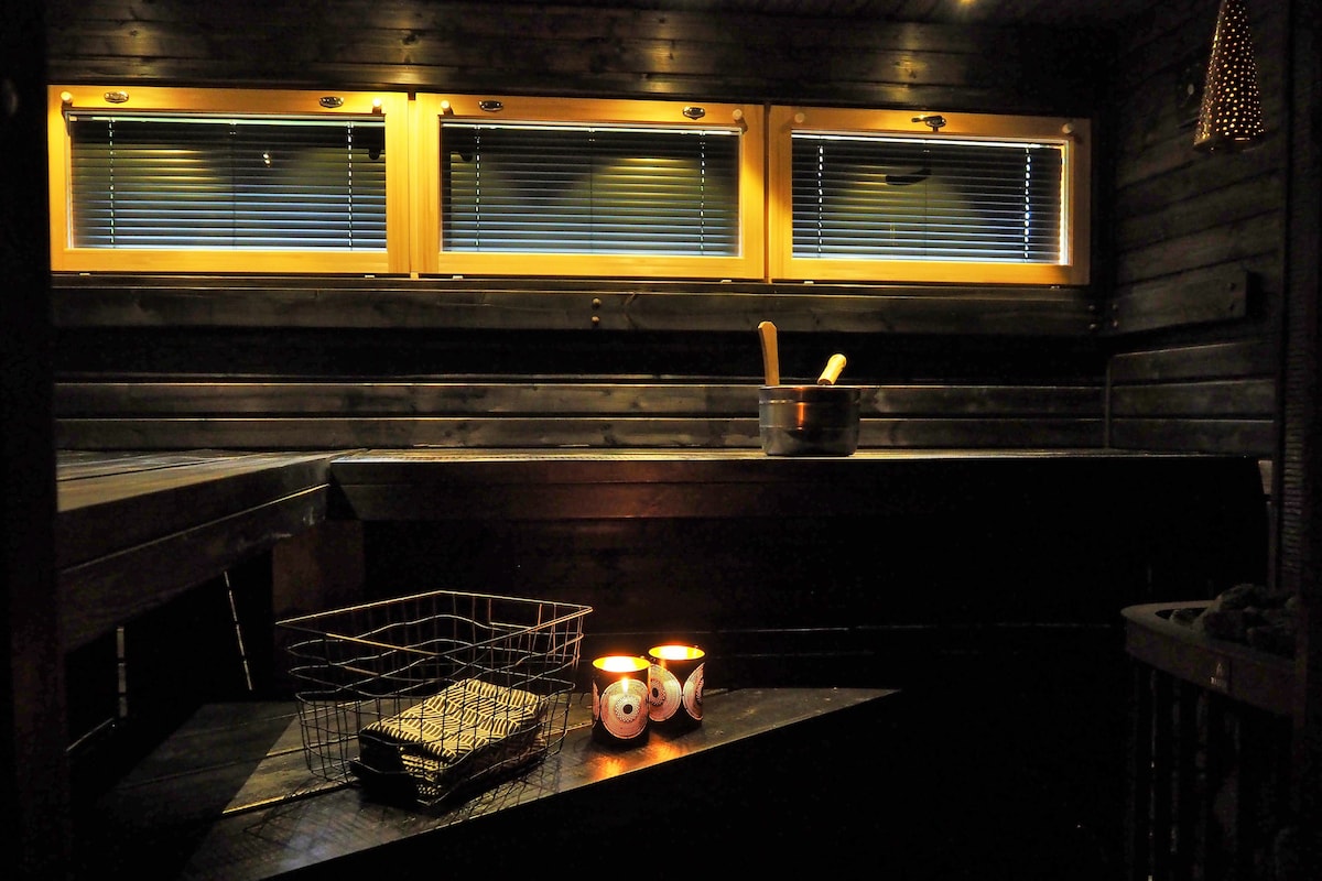 The interior of the sauna is crafted from dark wood, featuring two narrow windows that allow for soft natural light. A wooden bucket and ladle are positioned near a metal pot. Two candles provide a warm glow on a wooden shelf, enhancing the peaceful atmosphere.