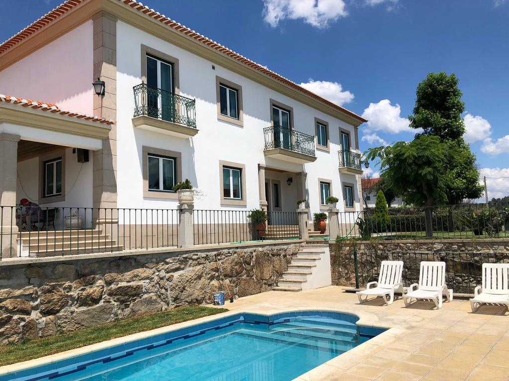 A two-story building with a white exterior features several balconies and large windows. The garden area includes a neatly designed pool surrounded by lounge chairs, with stone walls and landscaped greenery enhancing the outdoor space.