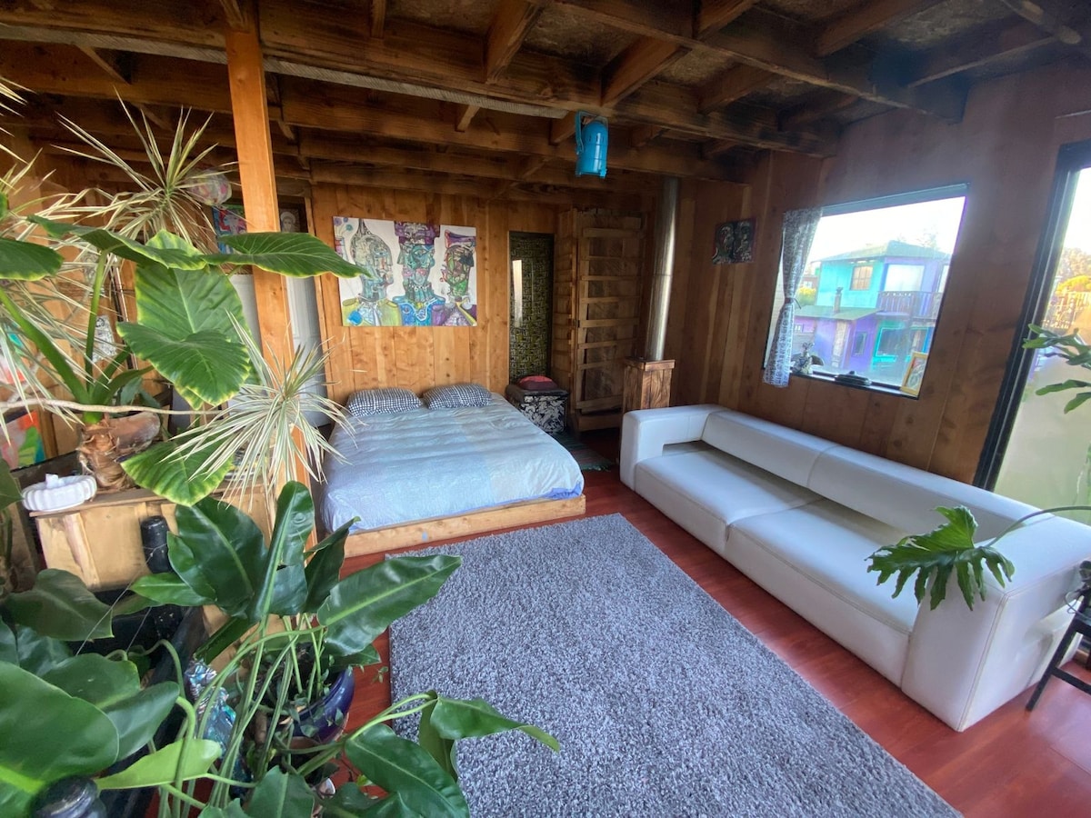 An open and airy sleeping area features natural wood accents and large windows that allow ample light. A spacious bed with soft bedding sits adjacent to a modern white sofa. Lush green plants add a touch of nature to the warm wooden interior.