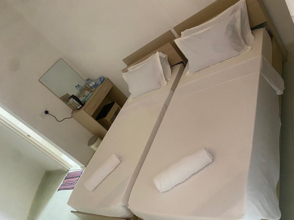 A room with twin beds is presented, each equipped with white linens and neatly rolled towels. A simple wooden desk and mirror are positioned nearby, while natural light enters through a partially open door, enhancing the bright and airy atmosphere.