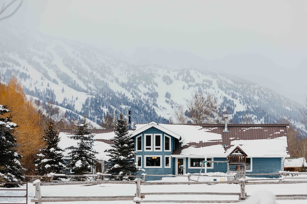 A snowy exterior of the home is set against a backdrop of snow-covered mountains. The house features a modern architectural design with large windows and a brown metal roof. Adjacent trees and a wooden fence frame the serene winter scene.