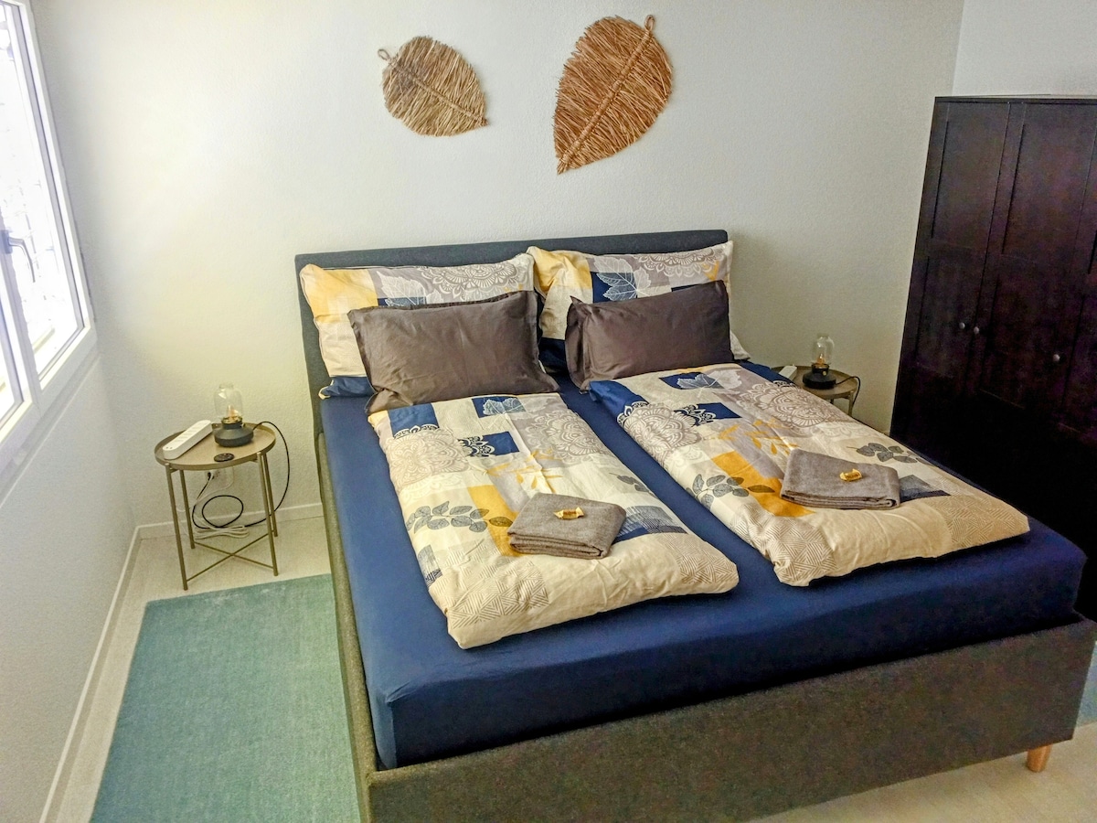 A comfortable sleeping area features a spacious bed with a blue fitted sheet and a quilted coverlet in a patchwork design. Two pillows are arranged at the headboard, and towels are neatly placed at the foot of the bed. Wall decor includes two woven leaf accents.