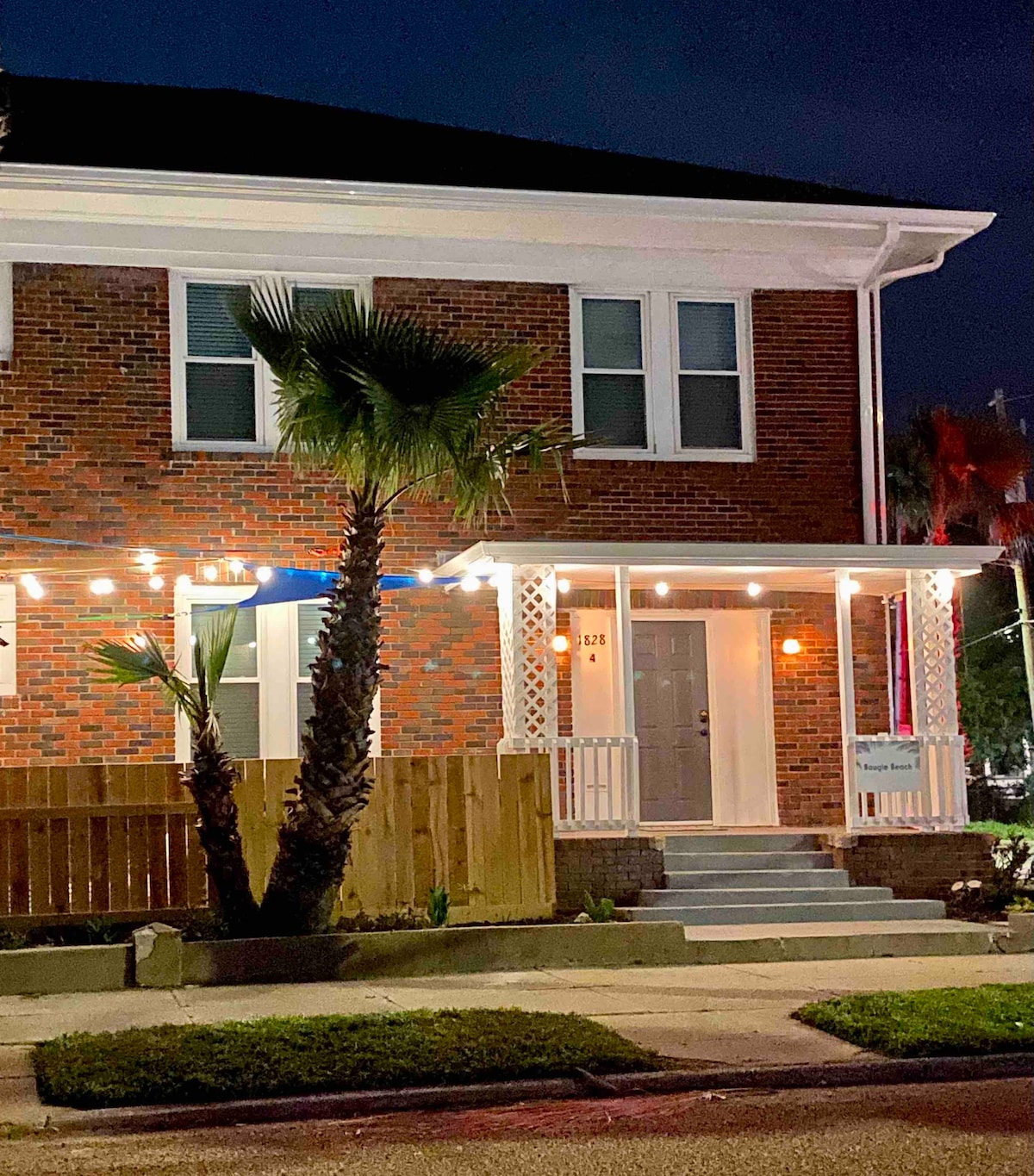 The exterior of a two-story brick condo is shown at night, illuminated by string lights adorning the porch. A tall palm tree enhances the tropical feel, while a wooden fence provides privacy. The entryway features a welcoming door framed by large windows.