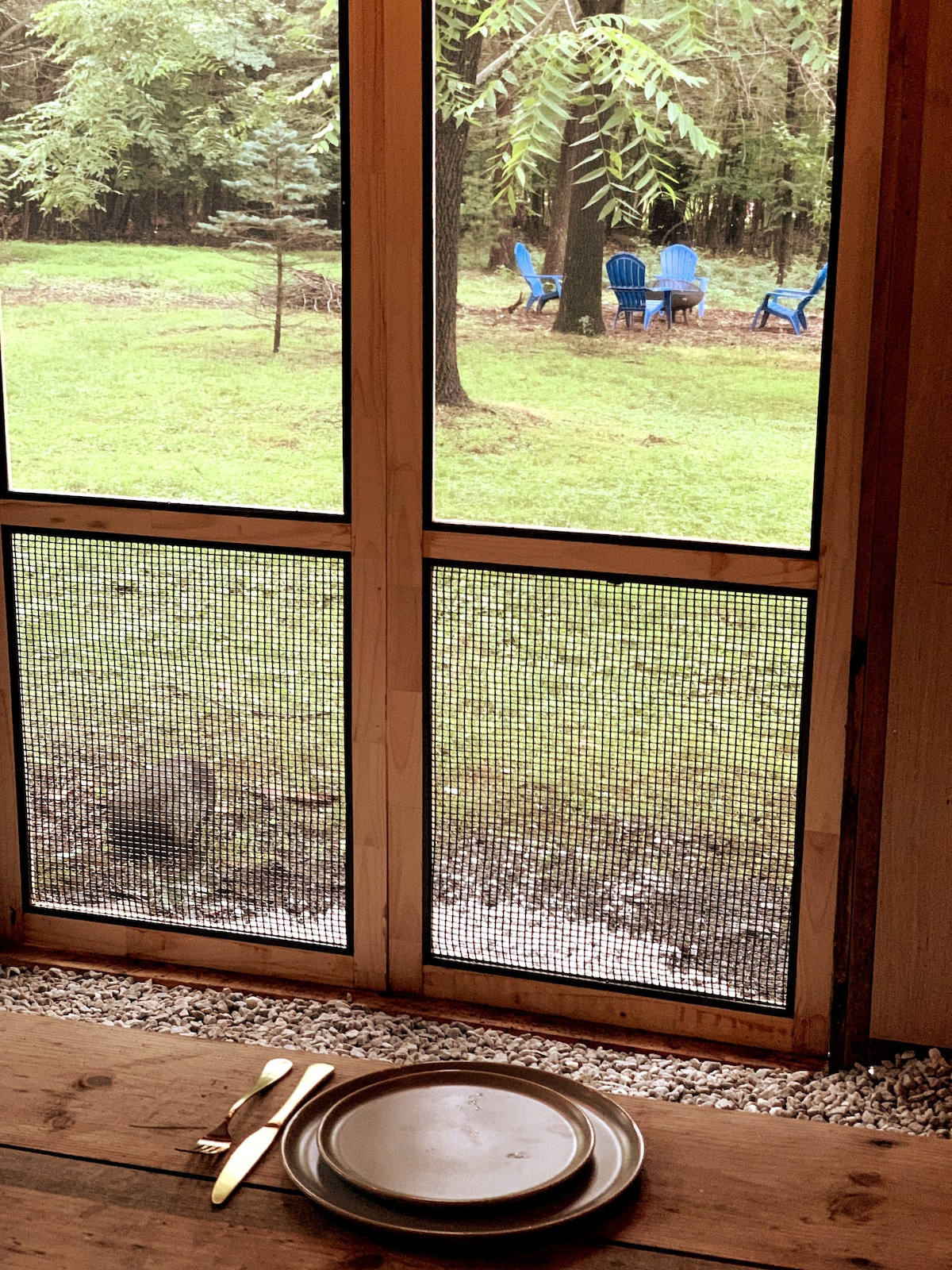 A view from inside the dining shed captures a table set with a plate and cutlery, framed by a screened window. Outside, blue Adirondack chairs are positioned on the grassy area among trees, providing a peaceful setting for relaxation.