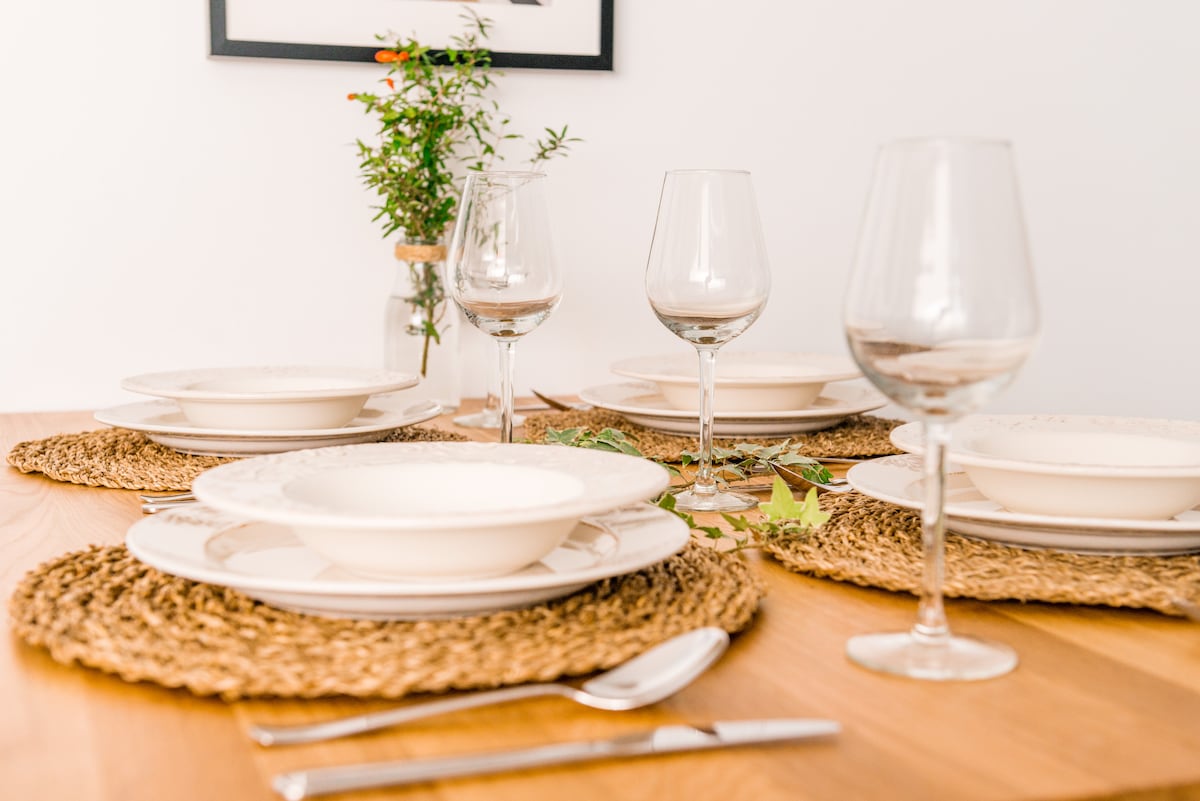 A dining table is set with four plates, accompanied by wine glasses and cutlery. Textured placemats add an organic touch. A small vase with greenery sits in the center, enhancing the table's simplicity and readiness for a meal.