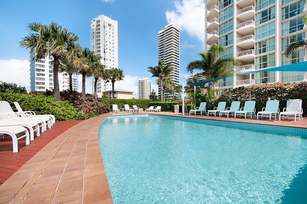 An inviting pool surrounded by a tiled deck features multiple lounge chairs positioned elegantly along its edge. Lush greenery and palm trees are visible, with tall buildings in the background, creating a relaxed resort atmosphere.