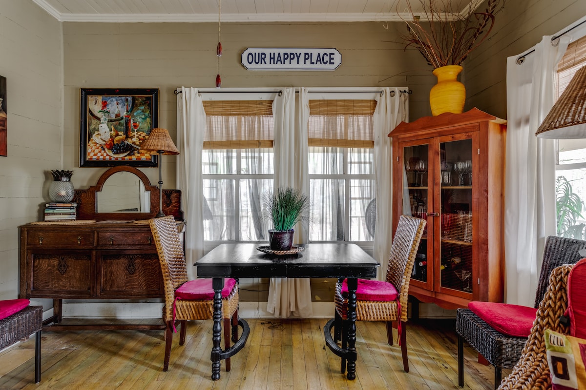 A cozy dining area features a dark wooden table surrounded by four chairs with colorful cushions. Natural light filters through the two windows, framed by sheer white curtains, while a glass-front cabinet displays decorative items. A potted plant centers the table, adding a touch of greenery.